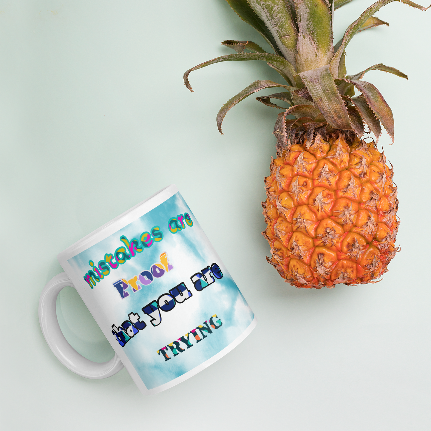 Ceramic mug with vibrant 'Mistakes are Proof' design next to a pineapple on a pastel background.