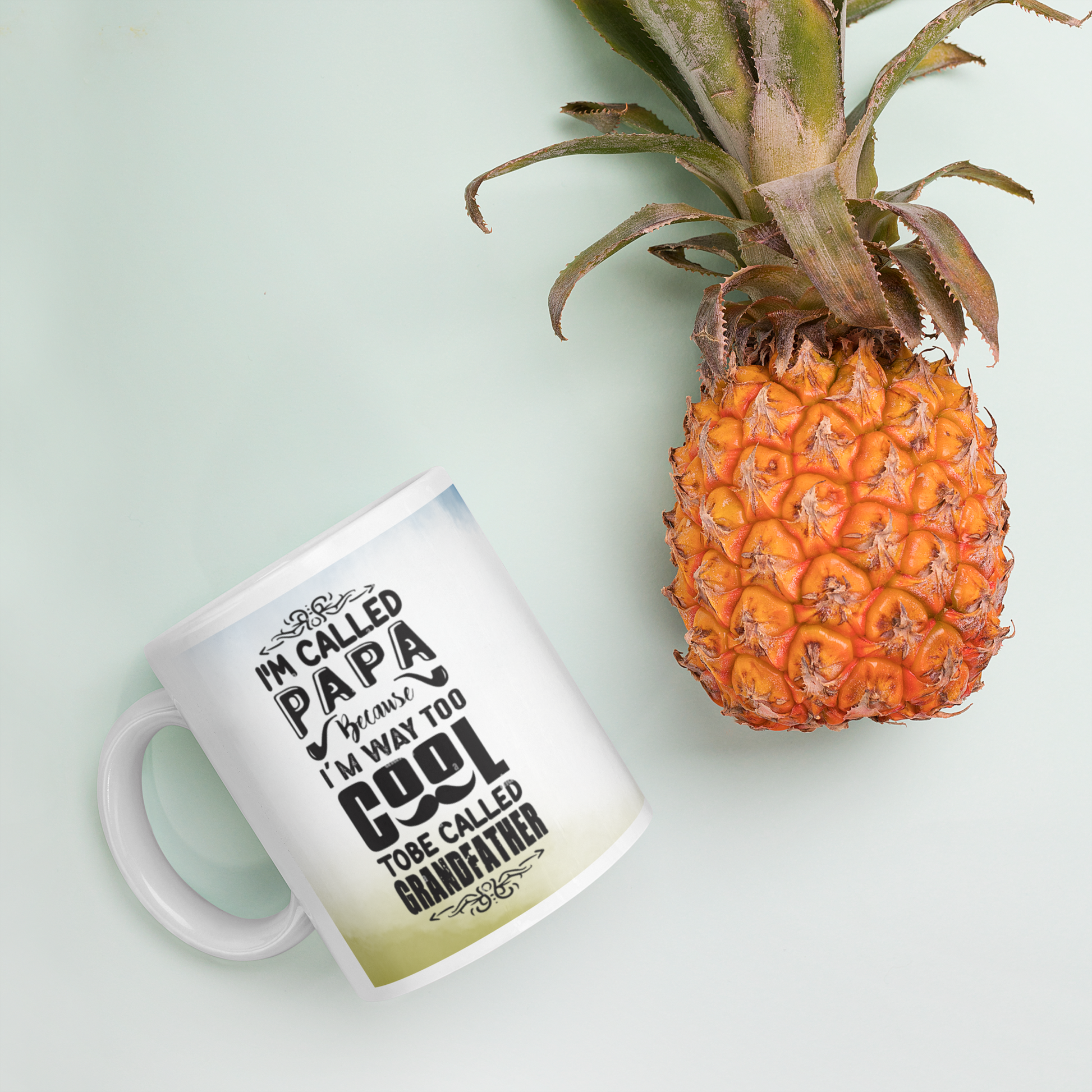 Ceramic mug with the text 'I'm called Papa, I'm way too cool to be called Grandfather' beside a fresh pineapple.